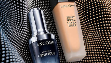Lancome Advanced Genifique serum and Teint Idole Ultra Wear foundation are shown against a dark, textured background with rows of glowing dots.