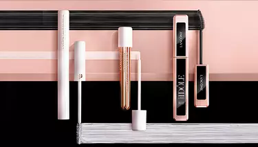 Various Lancome mascaras are displayed horizontally on a pink and black background with brush strokes, showing different packaging and brush types.