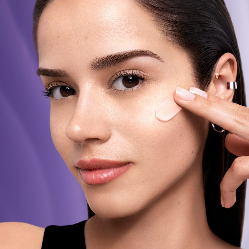 A young woman applies a skincare product to her cheek, showcasing her clear complexion and natural beauty.