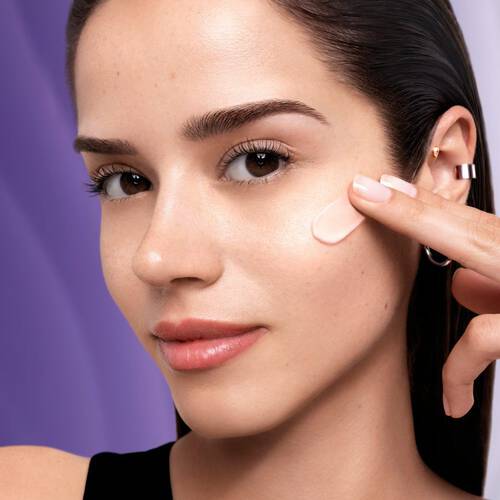 A young woman applies a skincare product to her cheek, showcasing her clear complexion and natural beauty.