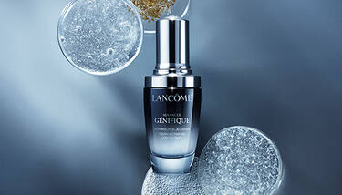 Lancome Advanced Genifique serum is centered, surrounded by petri dishes filled with various shimmering and granular substances, against a dark blue background.