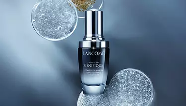 Lancome Advanced Genifique serum is centered, surrounded by petri dishes filled with various shimmering and granular substances, against a dark blue background.