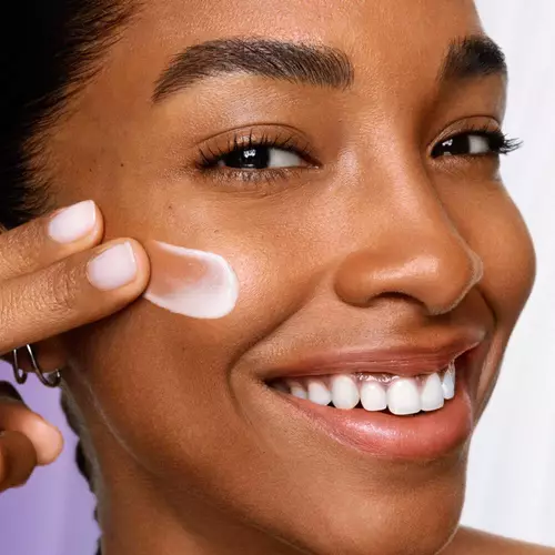 A smiling woman applies a creamy skincare product to her cheek, showcasing healthy, radiant skin.