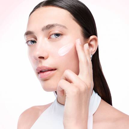 A woman with long, straight hair applying cream to her cheek against a soft pink background.