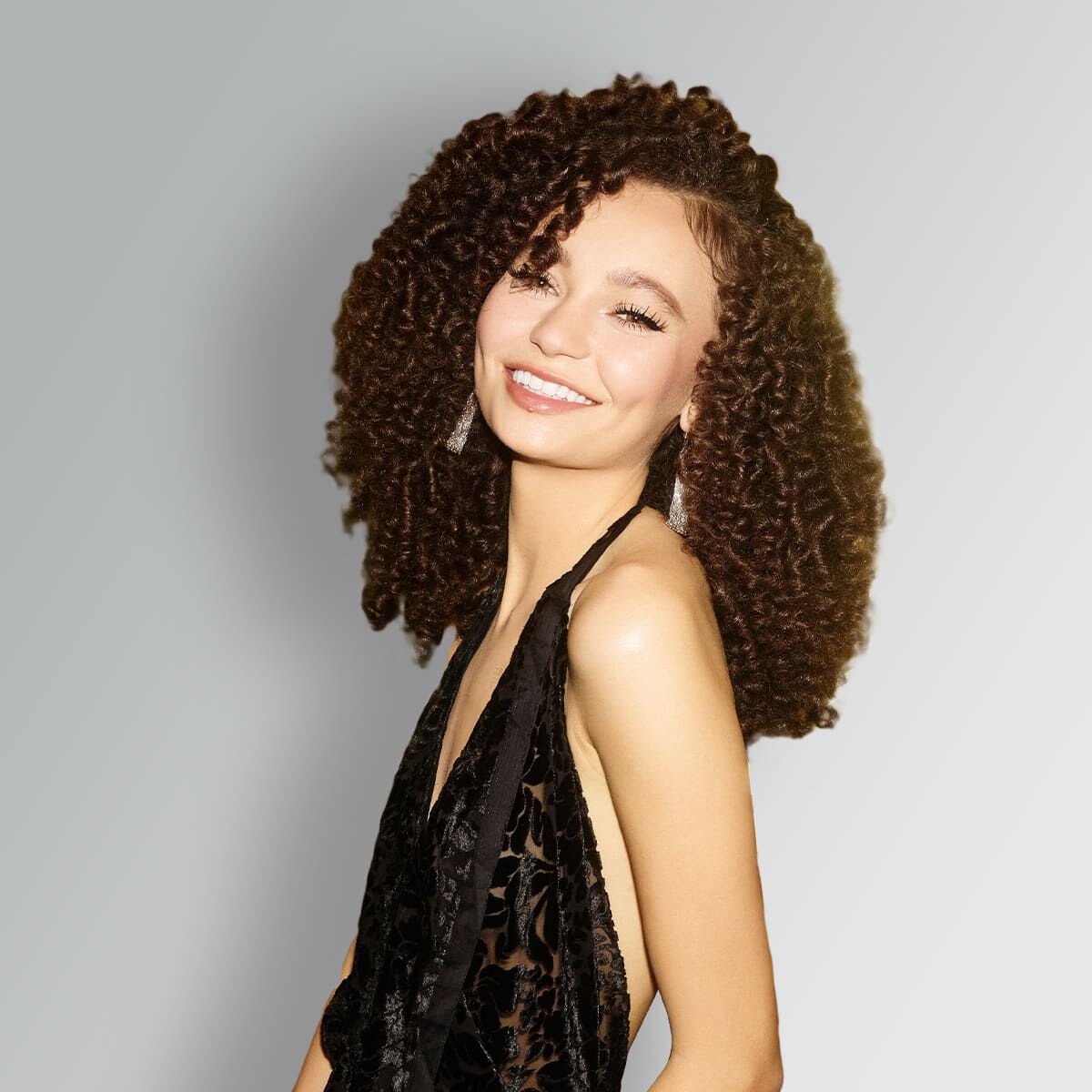 A smiling person with curly hair wearing a black outfit, posed against a light gray background.