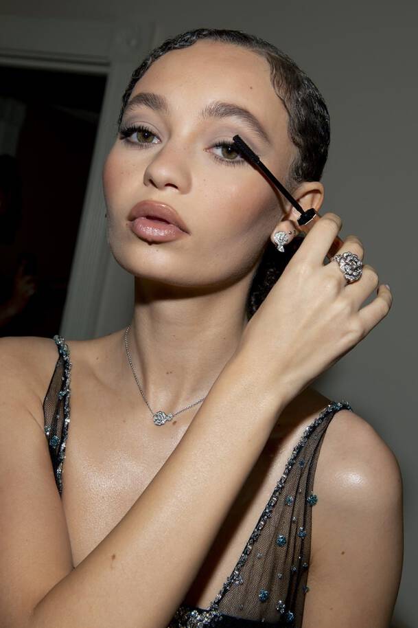 A woman with wet, slicked-back hair applies mascara, showcasing her makeup and jewelry while wearing a sleeveless, beaded dress.