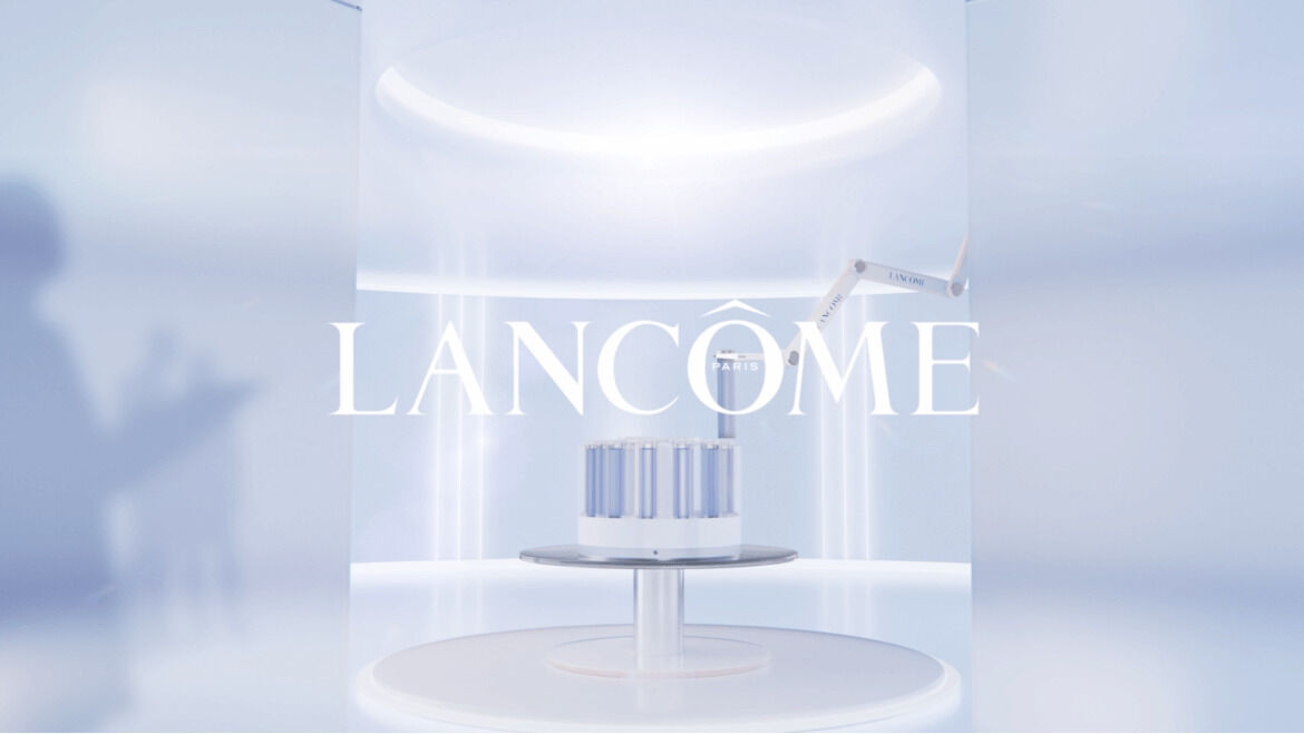 A sleek, modern display featuring the Lancome brand name prominently in the center, surrounded by a soft blue background and illuminated design. A robotic arm holds containers on a round white pedestal.