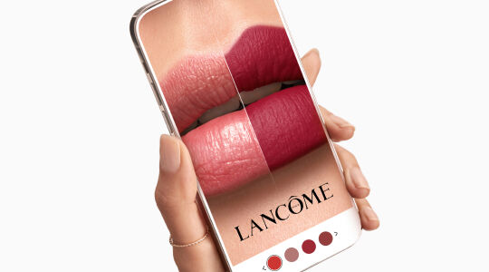 A hand holding a smartphone displaying a close-up of a model’s lips in four lipstick shades with the brand name Lancome at the bottom.