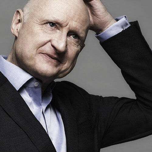 A man with a bald head and a thoughtful expression is tilting his head and scratching it, dressed in a dark suit with a light blue shirt.