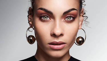 A close-up of a woman with vibrant green eyes and warm-toned eyeshadow, wearing distinctive gold hoop earrings with metallic spheres.