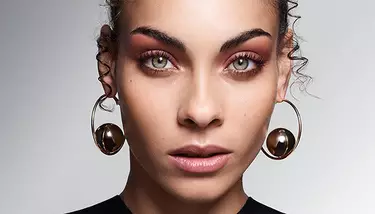 A close-up of a woman with vibrant green eyes and warm-toned eyeshadow, wearing distinctive gold hoop earrings with metallic spheres.