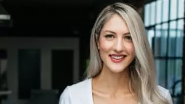 A smiling woman with long blonde hair and subtle makeup looks directly at the camera, against a blurred dark industrial background.