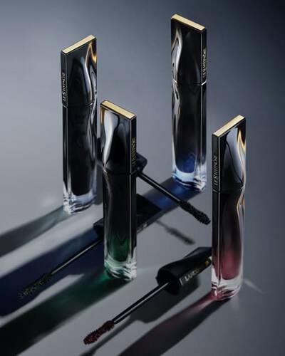 Four sleek, modern perfume bottles are arranged in a triangular formation on a dark surface, casting subtle shadows. The bottles have shiny metal caps and feature a gradient of colors from blue to green to red, enhancing their elegant appearance.