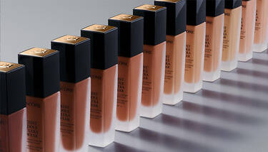 A diagonal line of Lancome Teint Idole Ultra Wear foundation bottles in varying shades, from dark to light, against a gray background.