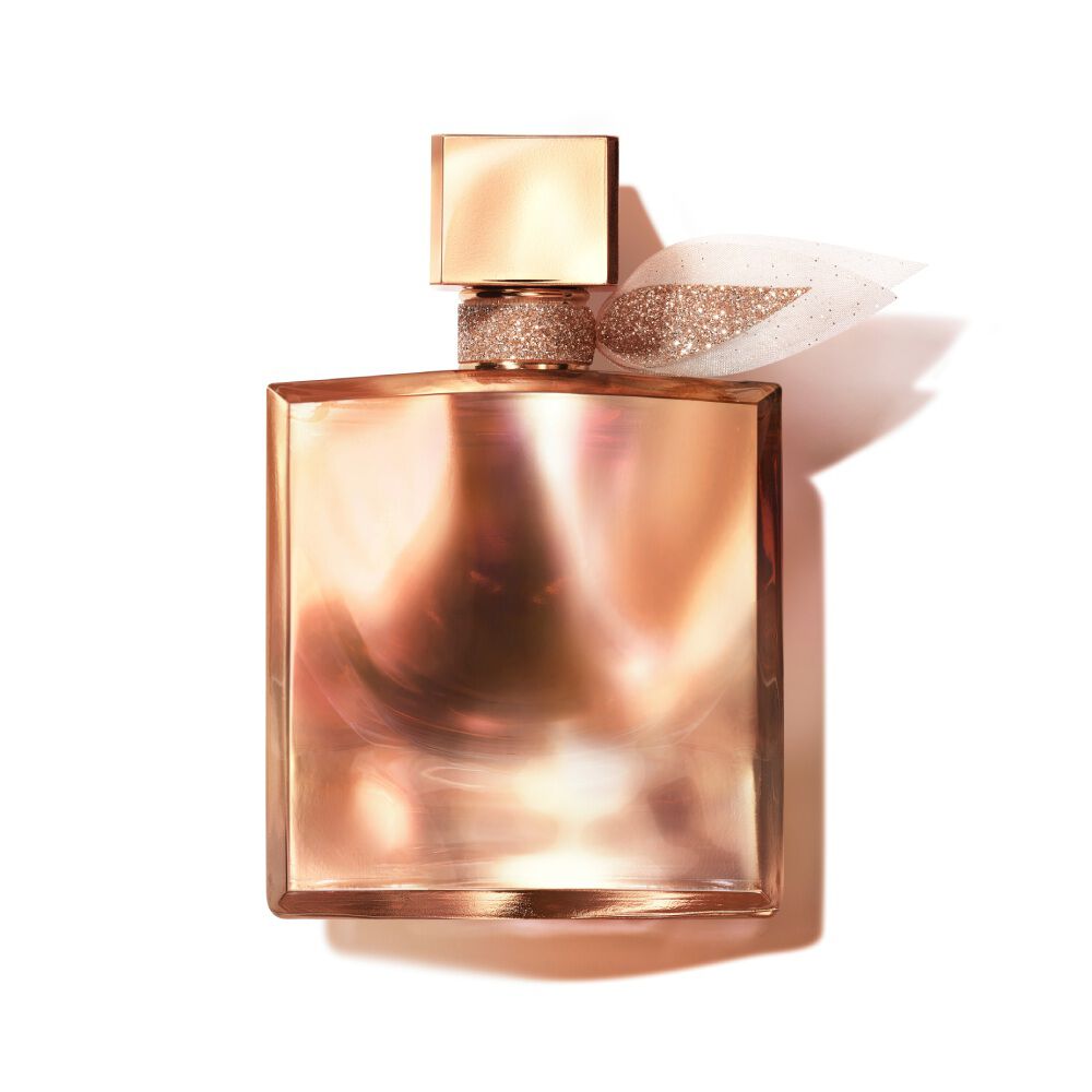A bottle of perfume with a square, elegant design, featuring a soft, shimmering rose gold hue and a decorative bow on the neck of the bottle.