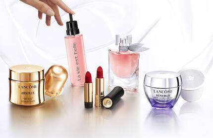 A collection of beauty products including skincare jars, lipsticks, and perfumes, arranged elegantly on a soft, light background. A hand is shown holding a pink spray bottle.