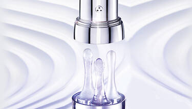 A close-up of a Lancome Genifique eye cream applicator with three pearl-shaped drops of product, set against a blurry white and purple background.
