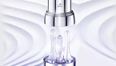 A close-up of a Lancome Genifique eye cream applicator with three pearl-shaped drops of product, set against a blurry white and purple background.