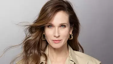 A woman with blue eyes and light makeup, wearing a beige collared shirt and gold abstract earrings, looks directly at the camera with her brown hair flowing around her shoulders.