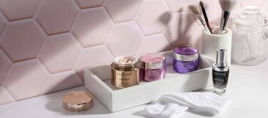 A collection of Lancome skincare products displayed on a white marble tray, including jars of cream and a serum, set against a light pink hexagonal tile background. Makeup brushes and a glass container are visible nearby, along with a white cloth.