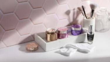A white tray holding several Lancome skincare jars and a serum bottle, along with cotton pads and makeup brushes, sits on a counter against a pink hexagonal tiled wall.