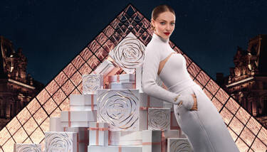 Amanda Seyfried, dressed elegantly in white, poses among a display of white Lancome gift boxes adorned with a rose design, set against the backdrop of the illuminated Louvre Pyramid at night.