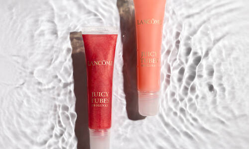 Two tubes of Lancome Juicy Tubes lip gloss sit on a textured surface that mimics rippling water. One tube contains a red gloss, and the other an orange gloss, both labeled "Juicy Tubes Original."
