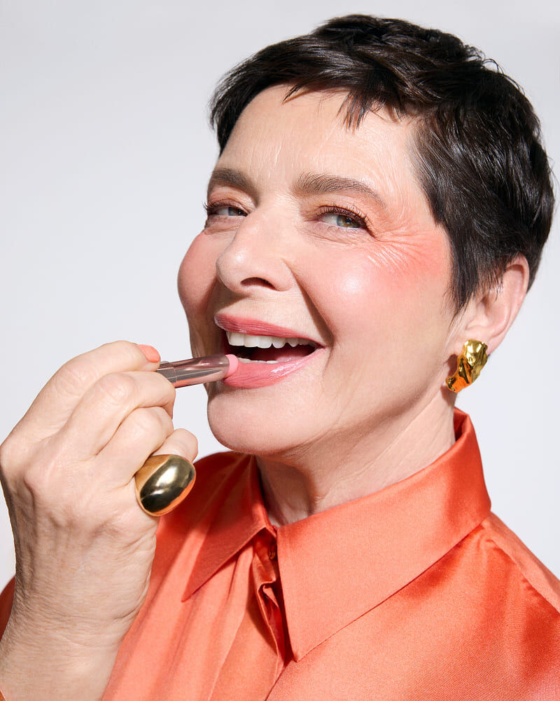 A smiling woman with short hair applies lipstick while wearing a bright orange blouse and bold jewelry.
