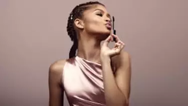 A woman in a satin top blows a kiss towards a mascara wand, with her braided hair styled back, against a neutral background.