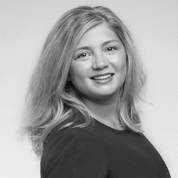 A woman with shoulder-length wavy hair smiles warmly, wearing a dark top against a light background.
