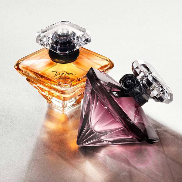 Two elegant perfume bottles are displayed on a textured surface. One bottle is an amber color with a square shape, while the other is a purple triangular bottle, both featuring distinctive crystal stoppers that catch the light.