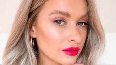 A close-up of a woman with platinum blonde hair, bright pink lipstick, winged eyeliner, and glowing cheeks, looking directly forward.