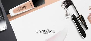 A flat lay of various Lancome skincare and makeup products, including a cream, serum, mascara, and fragrance, arranged aesthetically on a light background with the Lancome Paris logo prominently displayed.