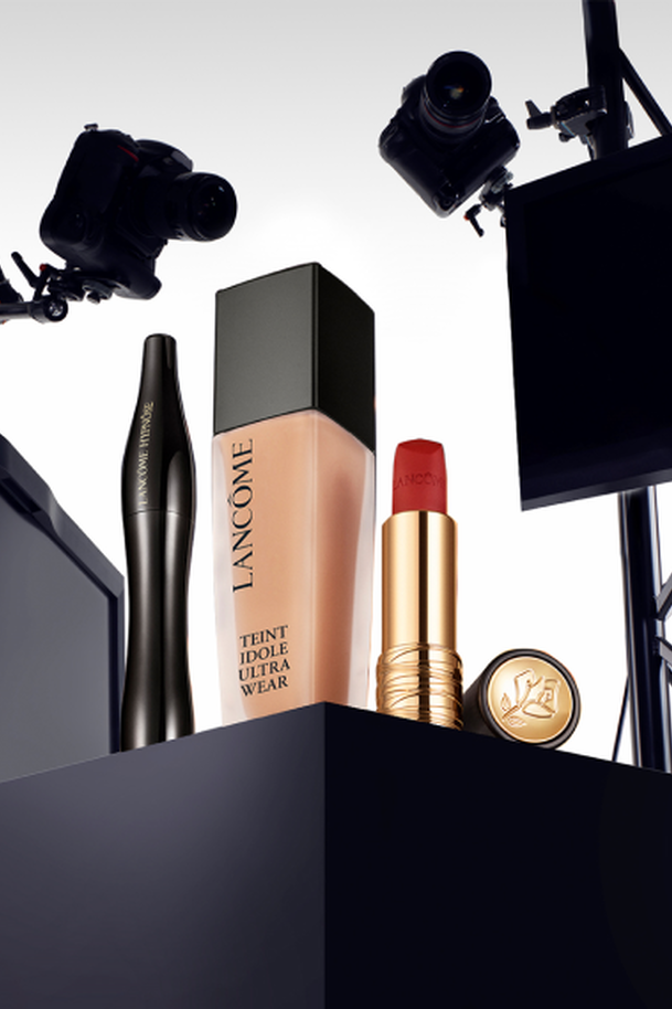 A display of Lancome cosmetics featuring a foundation bottle, a mascara, a lipstick tube, and a compact, all styled with cameras and lighting equipment in the background.