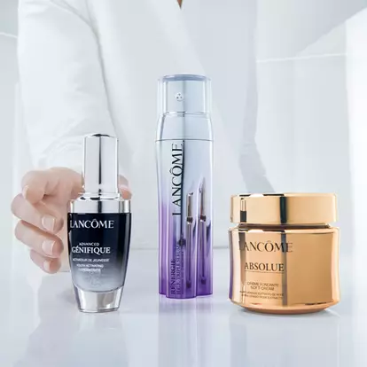A hand is reaching for three different luxury skincare products by Lancome, displayed on a minimalist glass surface.