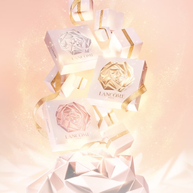 Three elegantly designed Lancôme gift boxes in pastel shades, surrounded by sparkling dust against a soft pink background.
