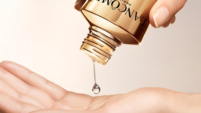 A hand holding a bottle of Lancôme serum, with a drop of the product about to fall into the palm.