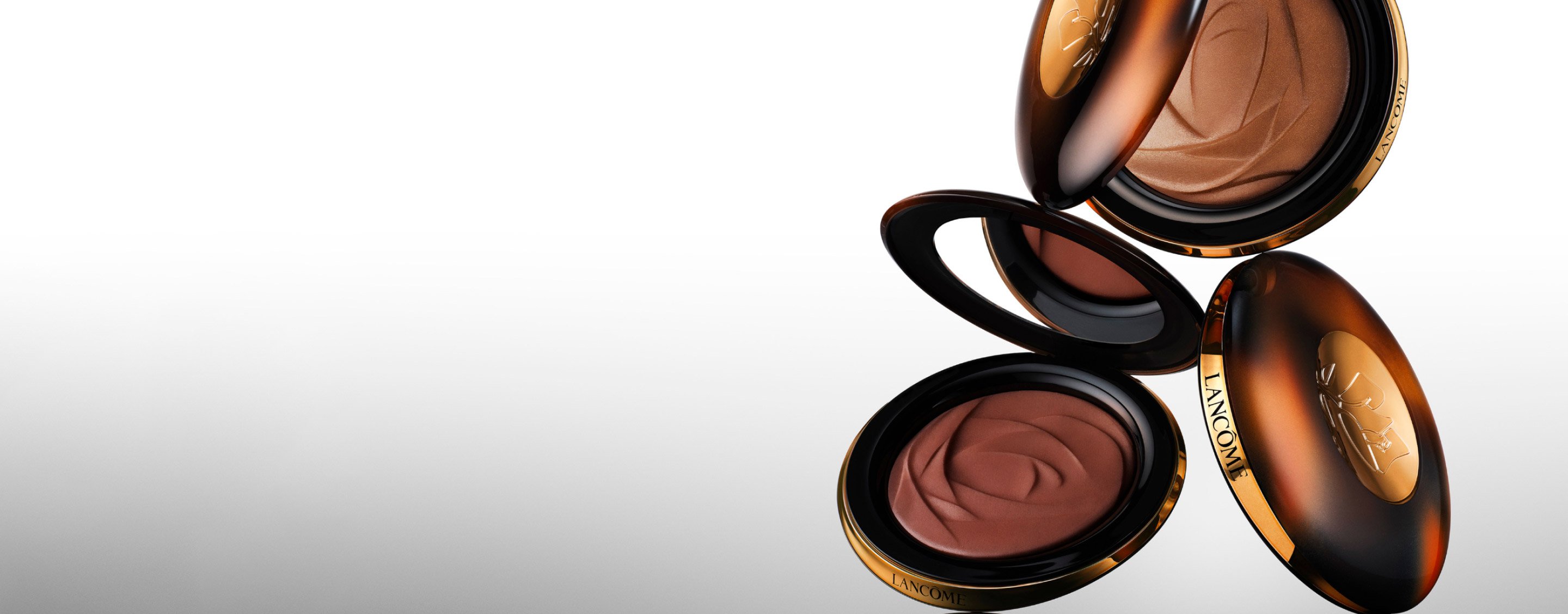Three Lancôme compact makeup cases in elegant oval shapes, featuring a gradient background that highlights their luxurious design.